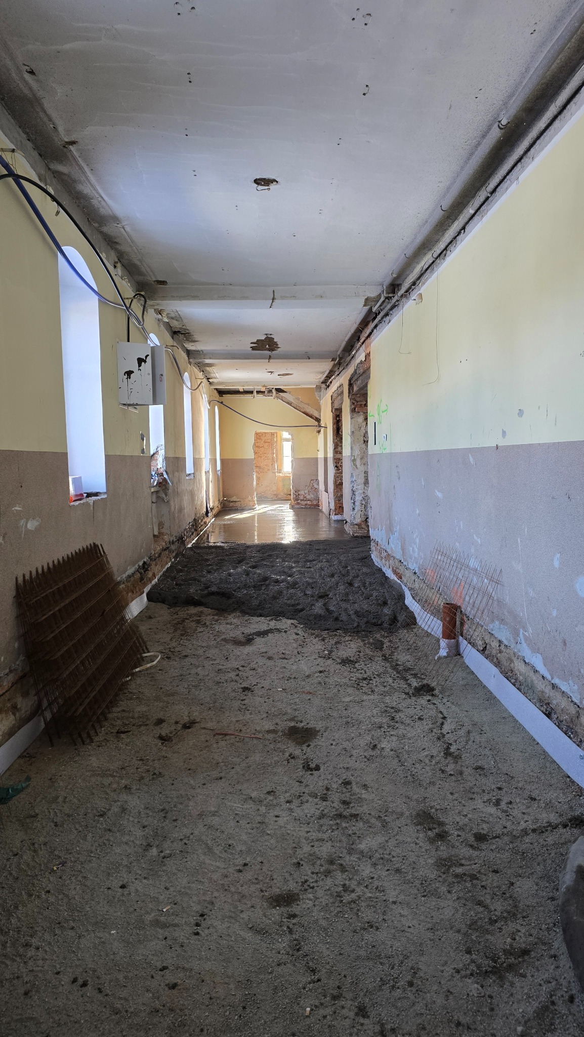 Wnętrze korytarza w trakcie wylewania posadzki. Widoczne okna, ściany w dwóch kolorach, częściowo wylana wylewka i siatka zbrojeniowa przy ścianie.