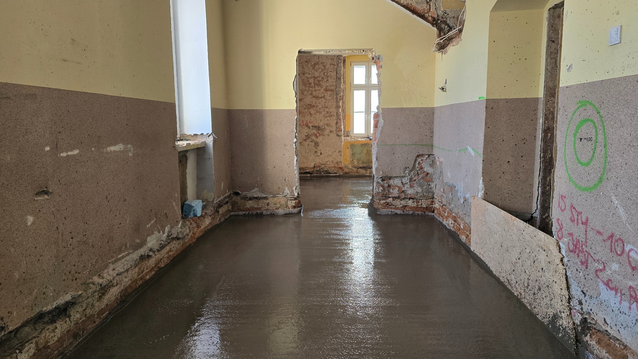 Świeża, szara wylewka podłogowa w remontowanym wnętrzu z odsłoniętą cegłą i zielonymi oznaczeniami na ścianie. Widok na sąsiednie pomieszczenia przez otwory drzwiowe.