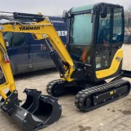 Żółto-czarna mini koparka Yanmar SV22 na gąsienicach, z łyżką i widocznym logo firmy, zaparkowana na kostce brukowej na tle niebieskiego samochodu dostawczego.