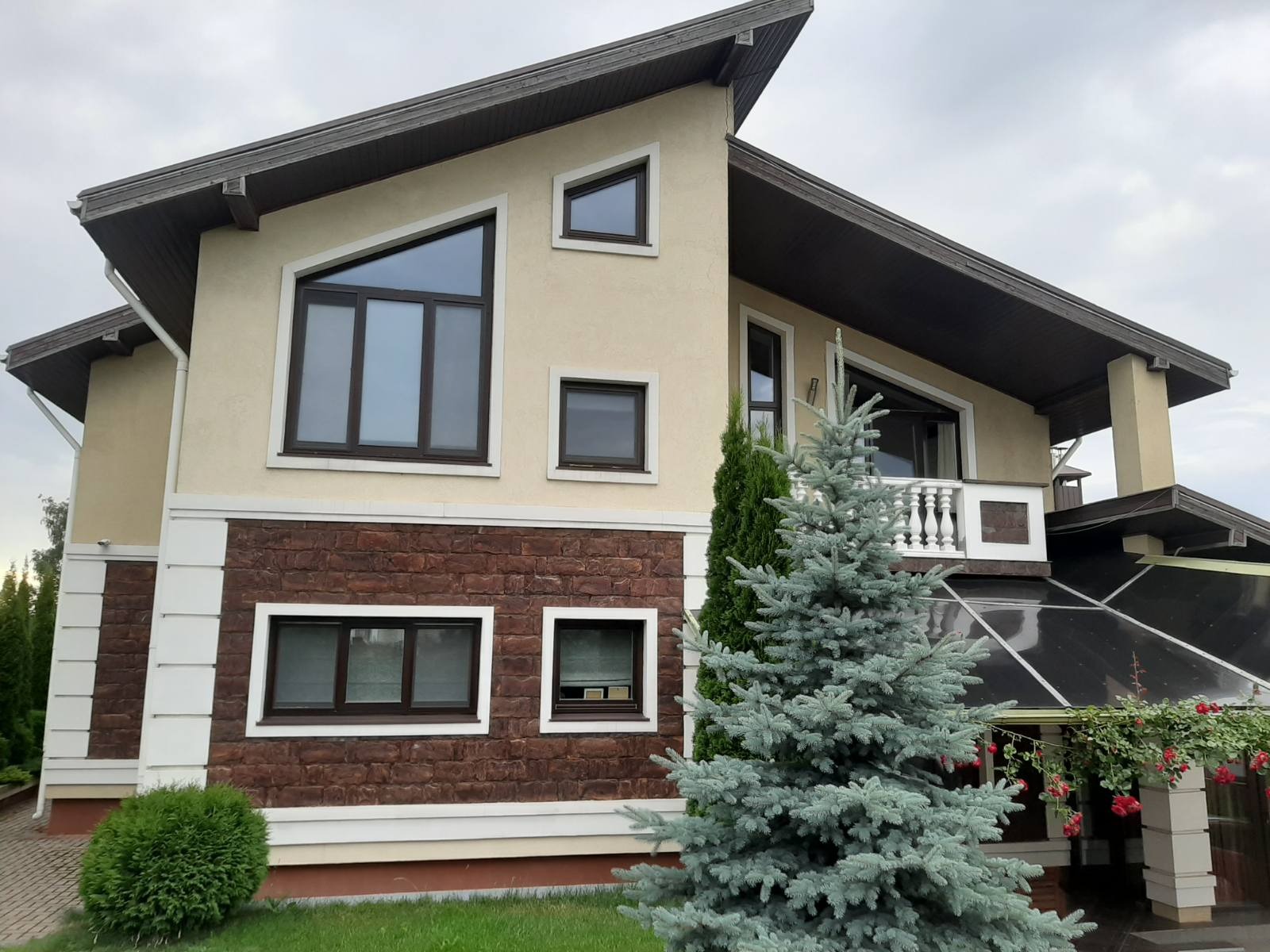 Elewacja dwukondygnacyjnego domu z brązowymi oknami, wykończona kamieniem i jasnym tynkiem, z dekoracyjną choinką na pierwszym planie.