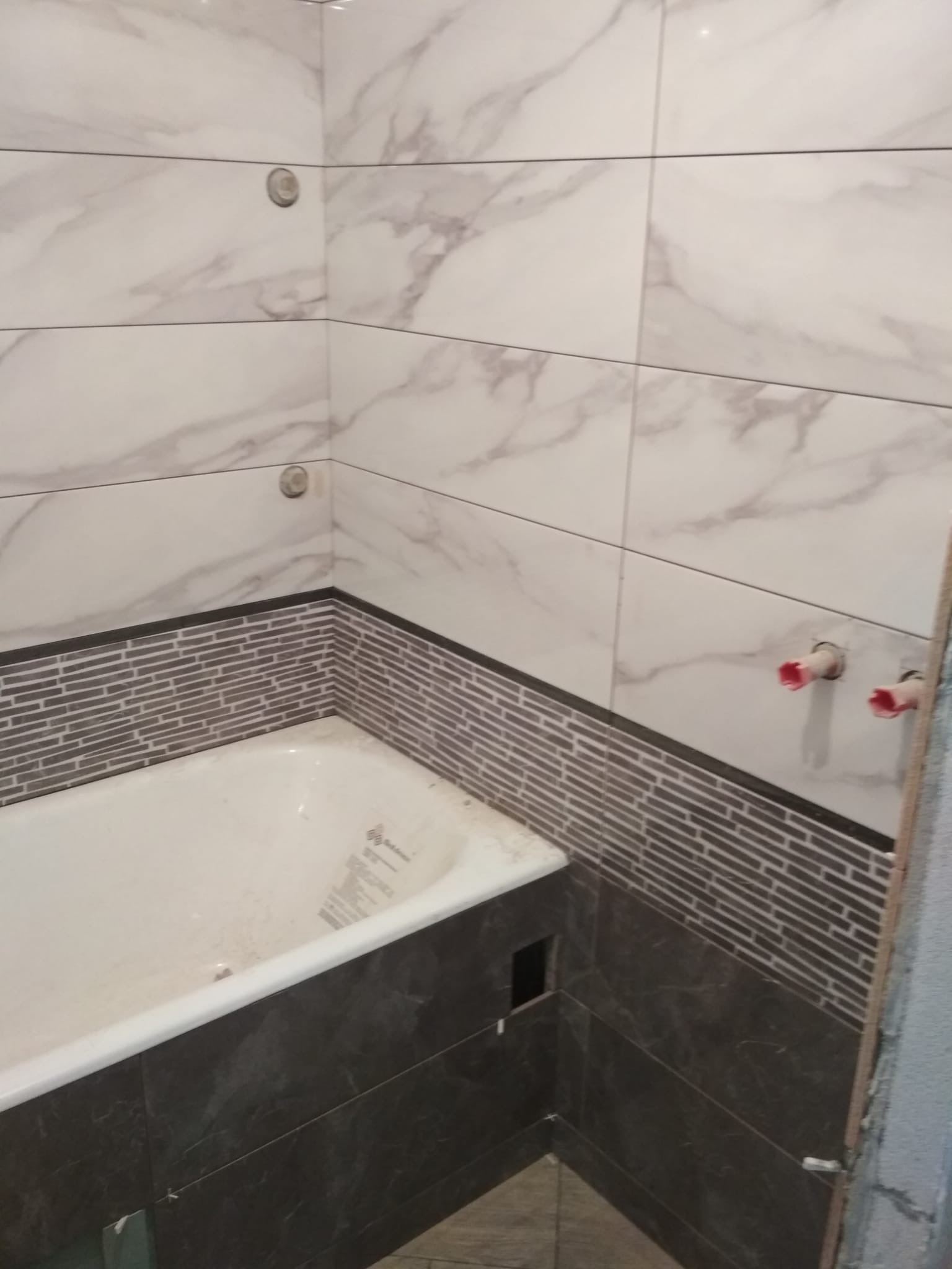 Wykończona obudowa wanny z ciemnymi płytkami imitującymi marmur, z mozaikowym pasem płytek nad wanną i białymi płytkami imitującymi marmur na ścianie, widoczne wystające rury instalacji wodnej.