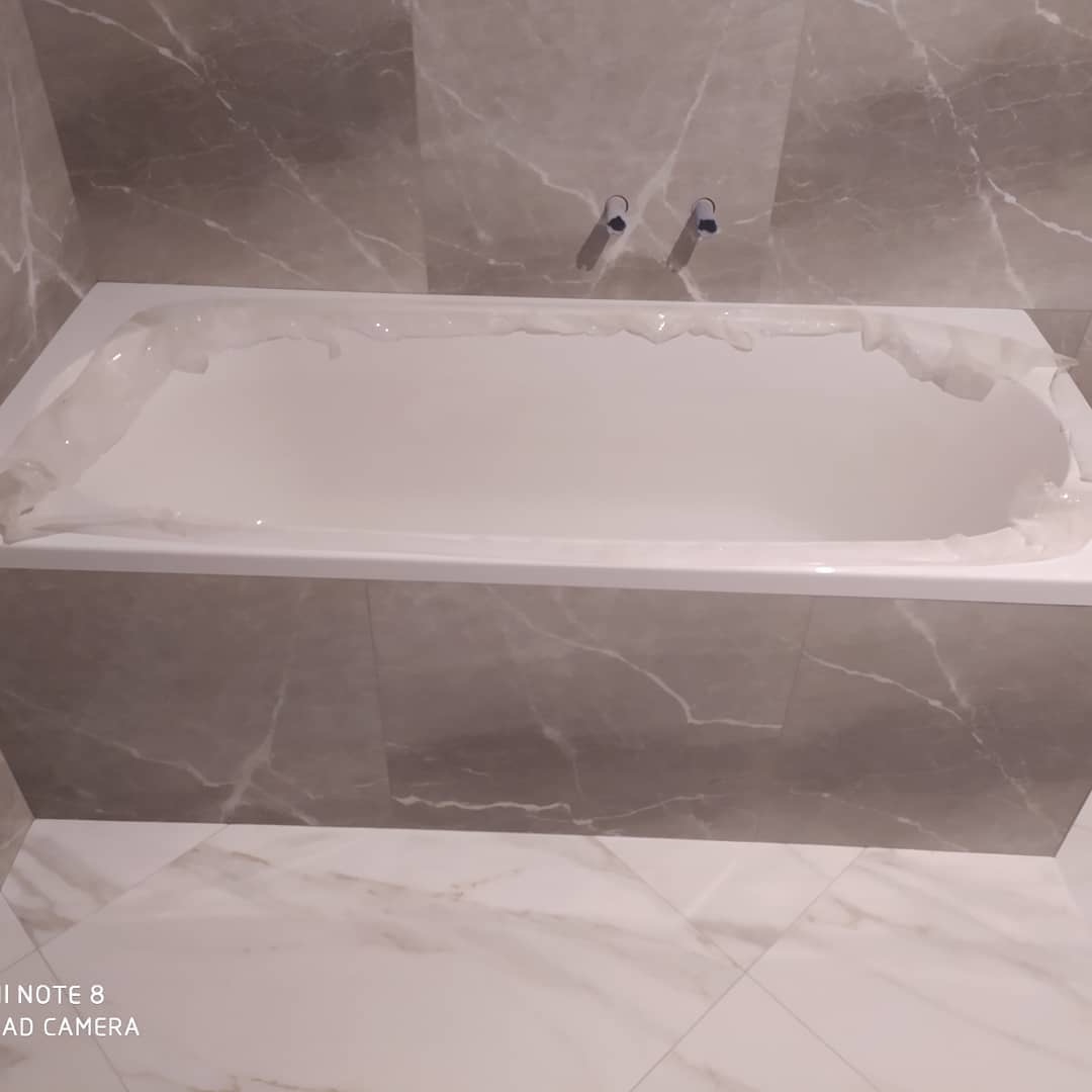 Biała wanna z zabezpieczającą folią, obłożona szarymi płytkami imitującymi marmur, z widocznymi chromowanymi bateriami na ścianie.