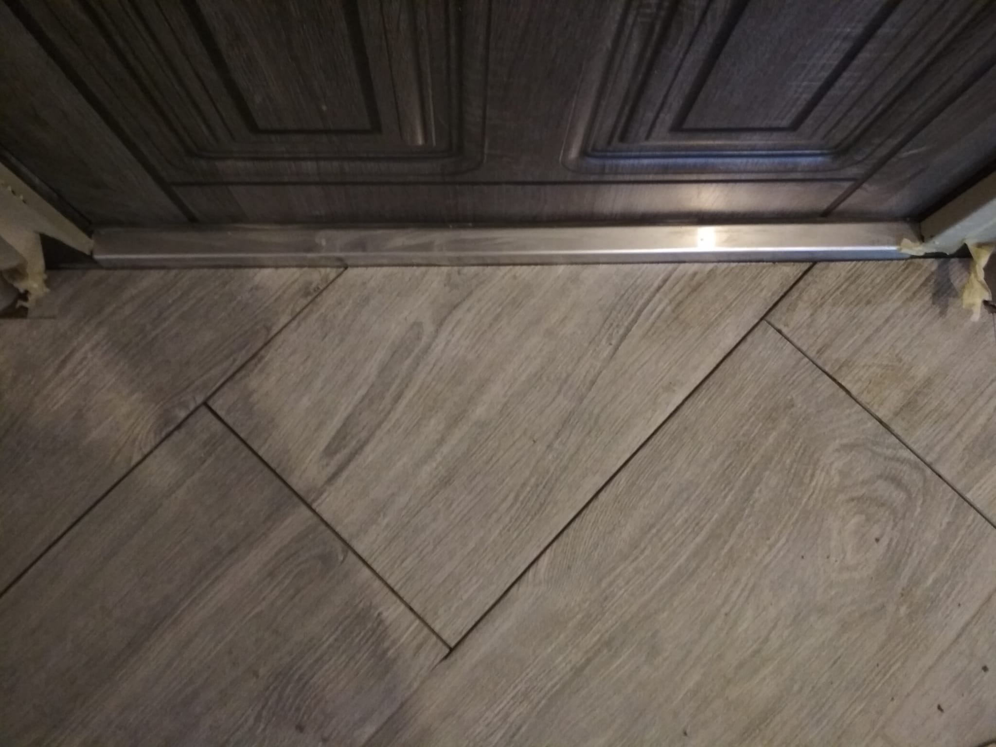 Szczegółowe ujęcie łączenia płytek podłogowych z drzwiami, widoczna metalowa listwa progowa i zabezpieczenia taśmą malarską.