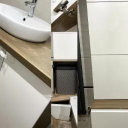 Nowoczesna łazienka z białą umywalką nablatową na blacie z drewnopodobnego laminatu, białe szafki z połyskiem, widoczna zawartość szafki z koszem na pranie i fragment kabiny prysznicowej.