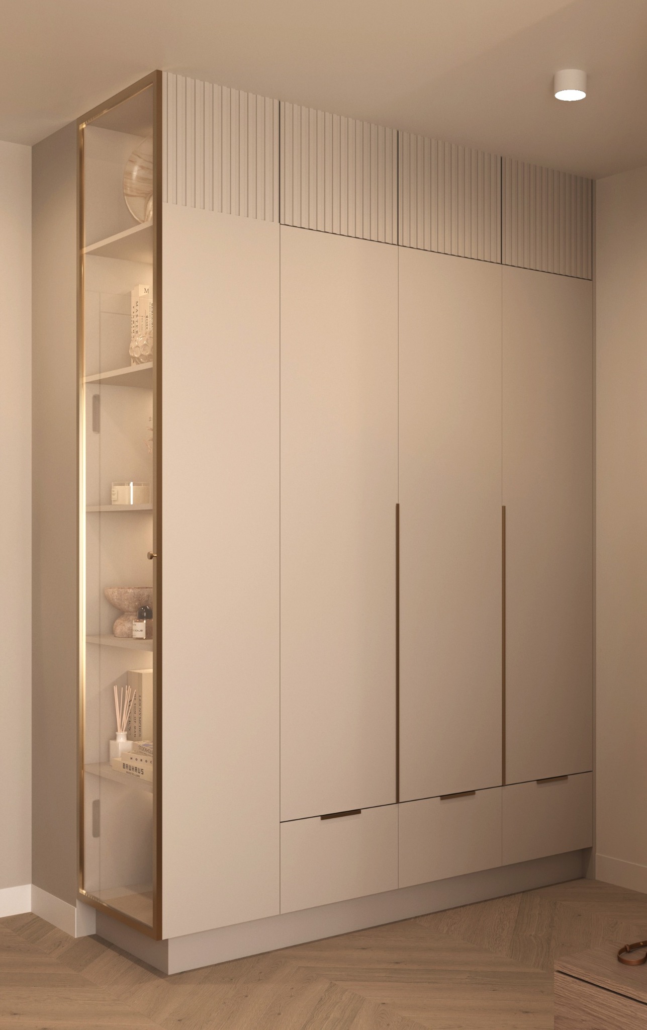 Wysoka, beżowa szafa wnękowa z przeszkloną witryną po lewej stronie, oświetloną od wewnątrz, eksponującą dekoracje i książki; nad szafą panel dekoracyjny z pionowymi żłobieniami.