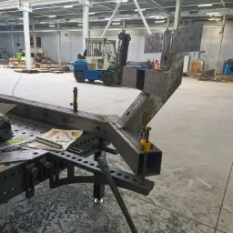 Spawana konstrukcja stalowa z blachy na stole spawalniczym w hali produkcyjnej, z widocznym wózkiem widłowym w tle. Detal wykonania konstrukcji.