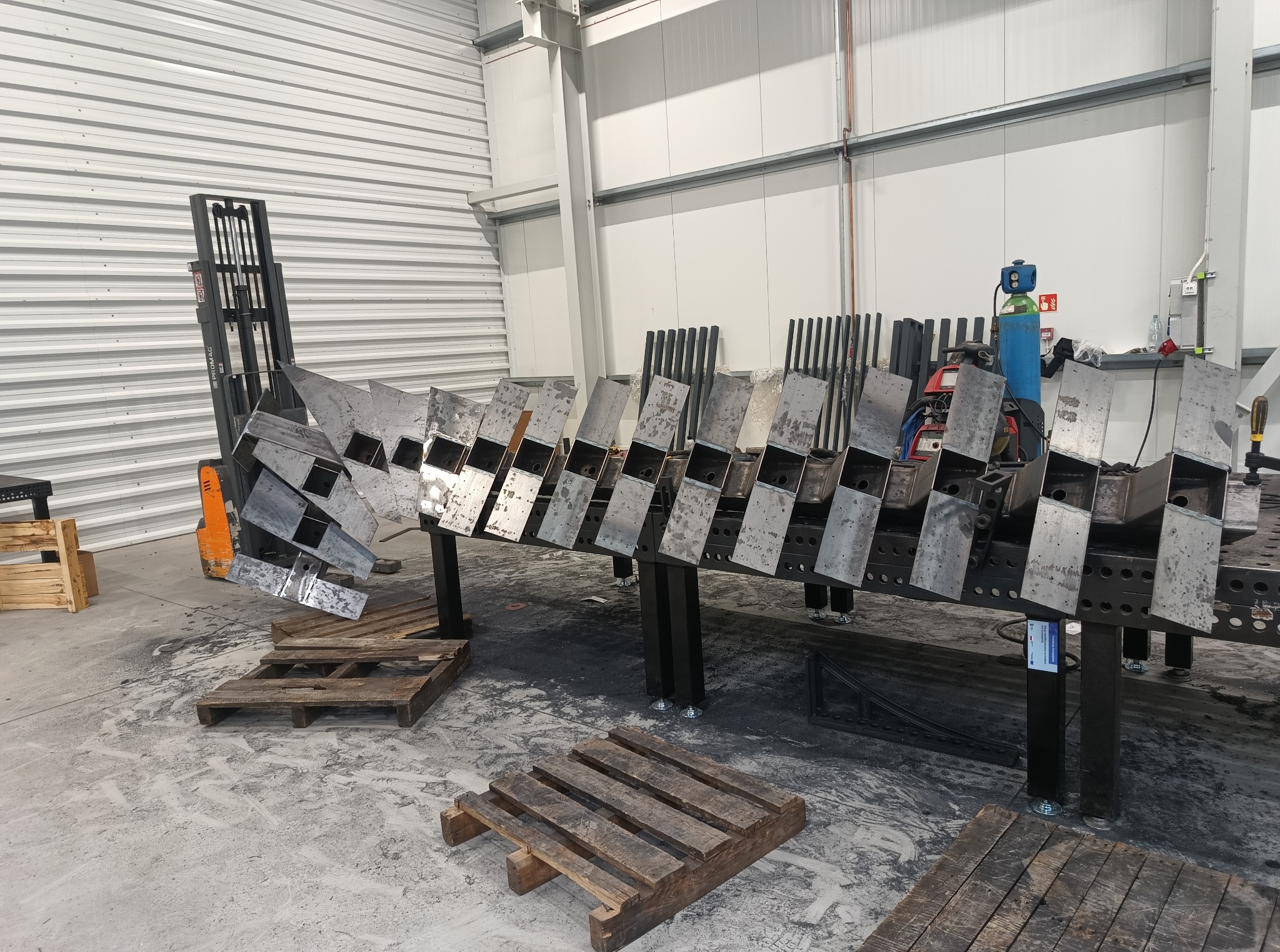 Warsztat ślusarski: spawana konstrukcja metalowa na stole, w tle wózek widłowy i narzędzia. Widoczne ślady pracy i palety.