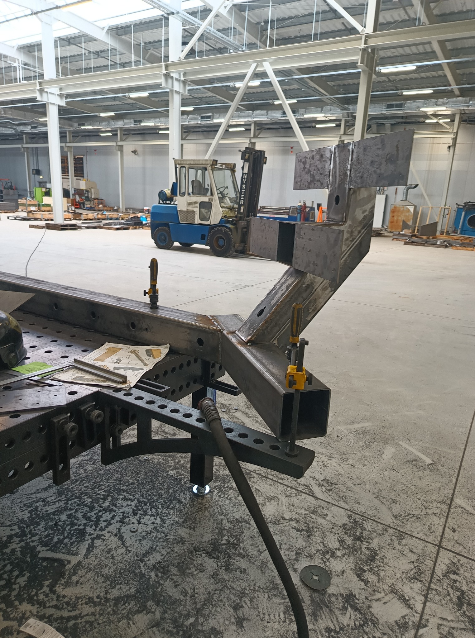 Spawana konstrukcja stalowa z blachy na stole spawalniczym w hali produkcyjnej, z widocznym wózkiem widłowym w tle. Detal wykonania konstrukcji.