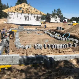 Budowa domu z betonu komórkowego: fundamenty w trakcie murowania, widoczne bloczki i pracujący murarze, w tle konstrukcja dachu i inne budynki.