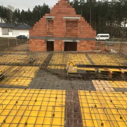 Wczesny etap budowy domu z czerwonej cegły, widok z góry na żółte szalunki i zbrojenie fundamentów, w tle las i biały bus.