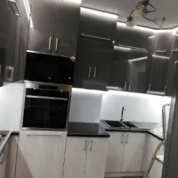 Nowoczesna kuchnia z szarymi, lakierowanymi szafkami górnymi i drewnopodobnymi szafkami dolnymi, podświetleniem LED podszafkowym i sufitowym, oraz drabina w rogu sugerująca prace wykończeniowe.