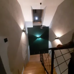 Klatka schodowa po remoncie: drewniane schody, szare ściany z nowoczesnymi kinkietami i czarną ścianą akcentującą, widoczne ślady szpachlowania na ścianach.