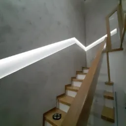 Klatka schodowa z drewnianymi schodami, szarą ścianą wykończoną efektem betonu, podświetleniem LED wzdłuż ściany i szklaną balustradą.