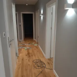 Korytarz po odświeżeniu: szare ściany, drewnopodobna podłoga, białe drzwi i listwy przypodłogowe, nowoczesne oświetlenie ścienne. Na podłodze widoczne ślady po remoncie: resztki taśmy malarskiej...