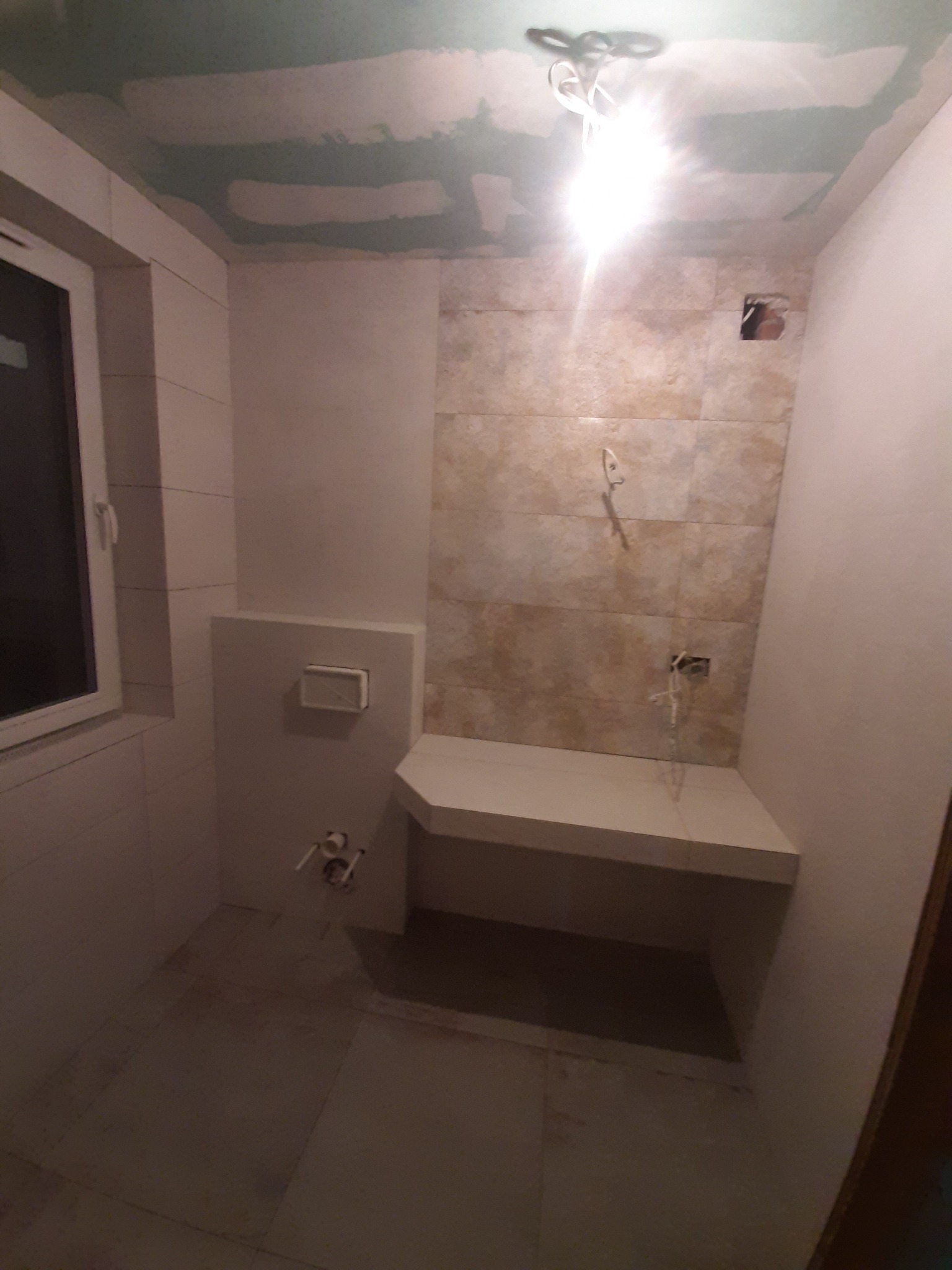 Wnętrze łazienki w trakcie remontu: położone płytki na ścianie, częściowo wykończona zabudowa podtynkowego WC, surowy sufit z widocznymi łączeniami płyt gipsowo-kartonowych.