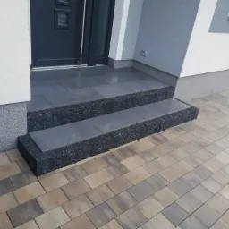 Schody wejściowe do domu wyłożone szarą kostką brukową, z ciemnym, ziarnistym obramowaniem, prowadzące do drzwi wejściowych; obok ułożona kostka brukowa w odcieniach beżu i szarości.