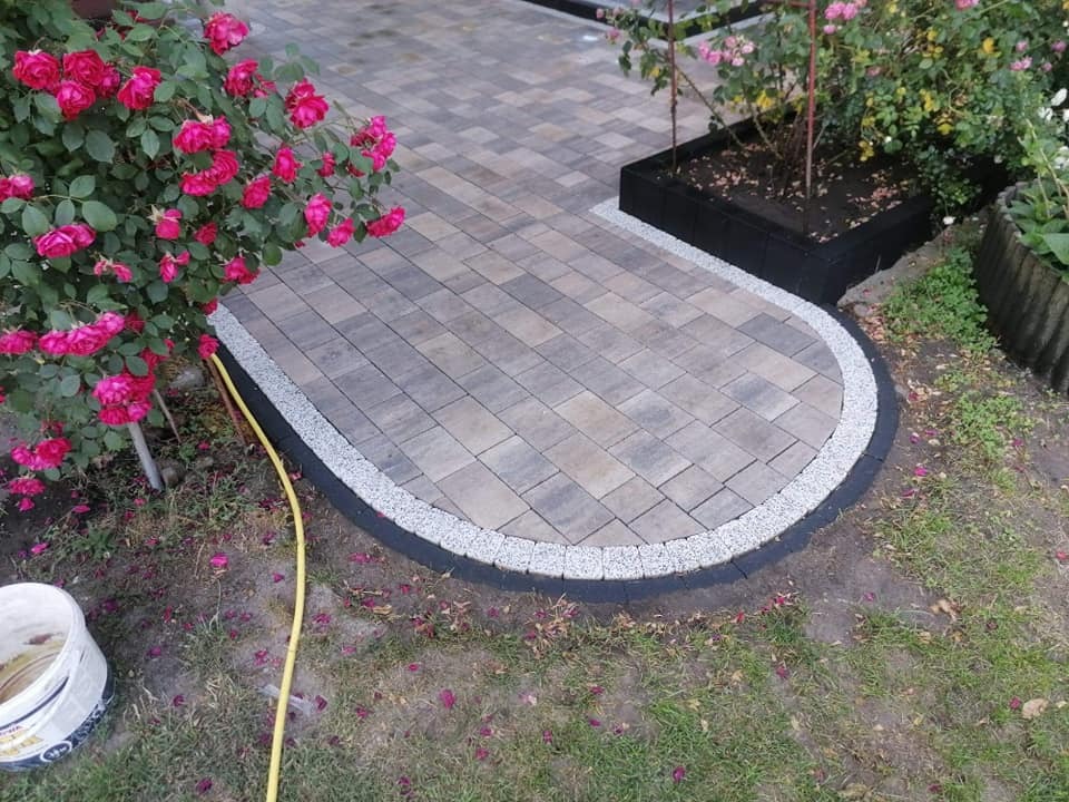 Ułożona kostka brukowa w odcieniach szarości z obrzeżem z drobnych kamieni, tworząca półokrągłą ścieżkę przy rabacie z kwitnącymi różami; widoczny biały kubeł i żółty wąż ogrodowy.