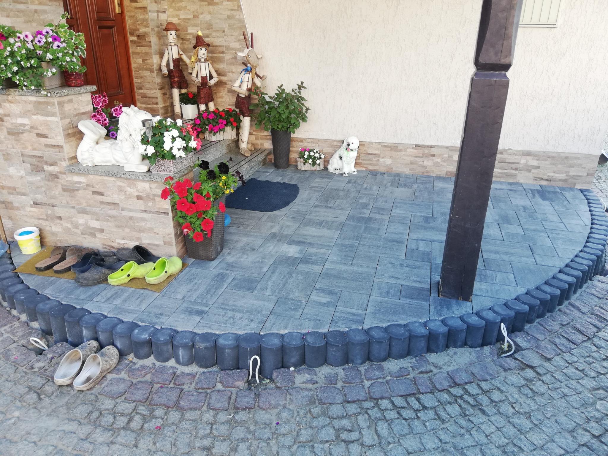 Ułożona szara kostka brukowa na podwórku z dekoracjami: figurki, donice z kwiatami, rzeźba lwa i zabawka pies.