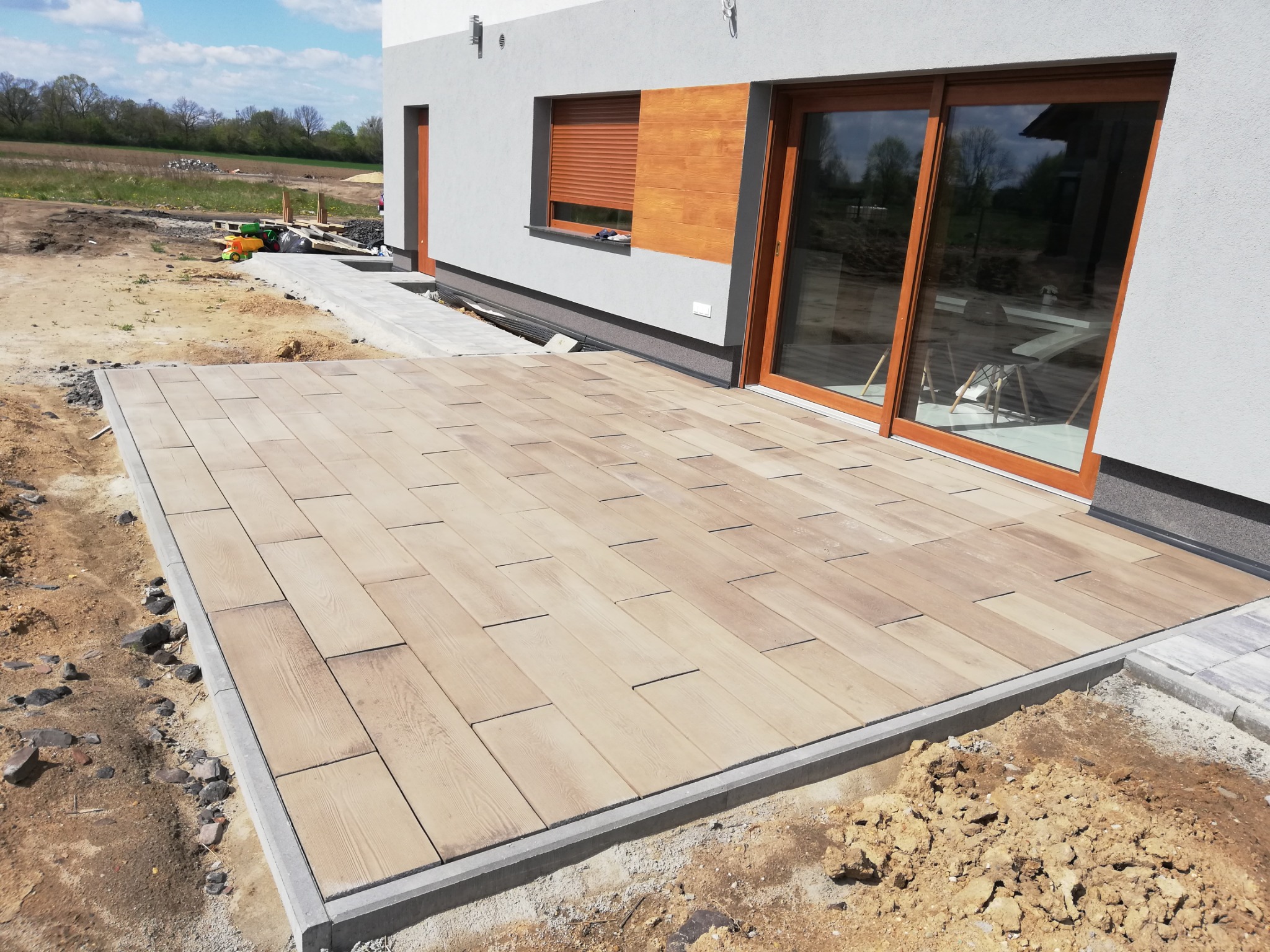 Nowoczesny taras z jasnej kostki brukowej imitującej drewno, obrzeża betonowe, widok na dom z dużymi oknami przesuwnymi i częściowo zagospodarowany teren.