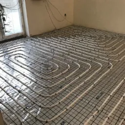 Białe rury ogrzewania podłogowego ułożone spiralnie na folii z nadrukiem 'KELLER' w surowym pomieszczeniu z oknem i wystającymi kablami.