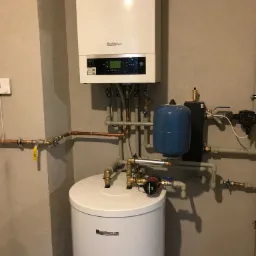 Biały piec gazowy Buderus zamontowany na ścianie, połączony rurami miedzianymi i plastikowymi z białym zasobnikiem ciepłej wody i niebieskim naczyniem przeponowym, widoczne zawory i pompa obiegowa...