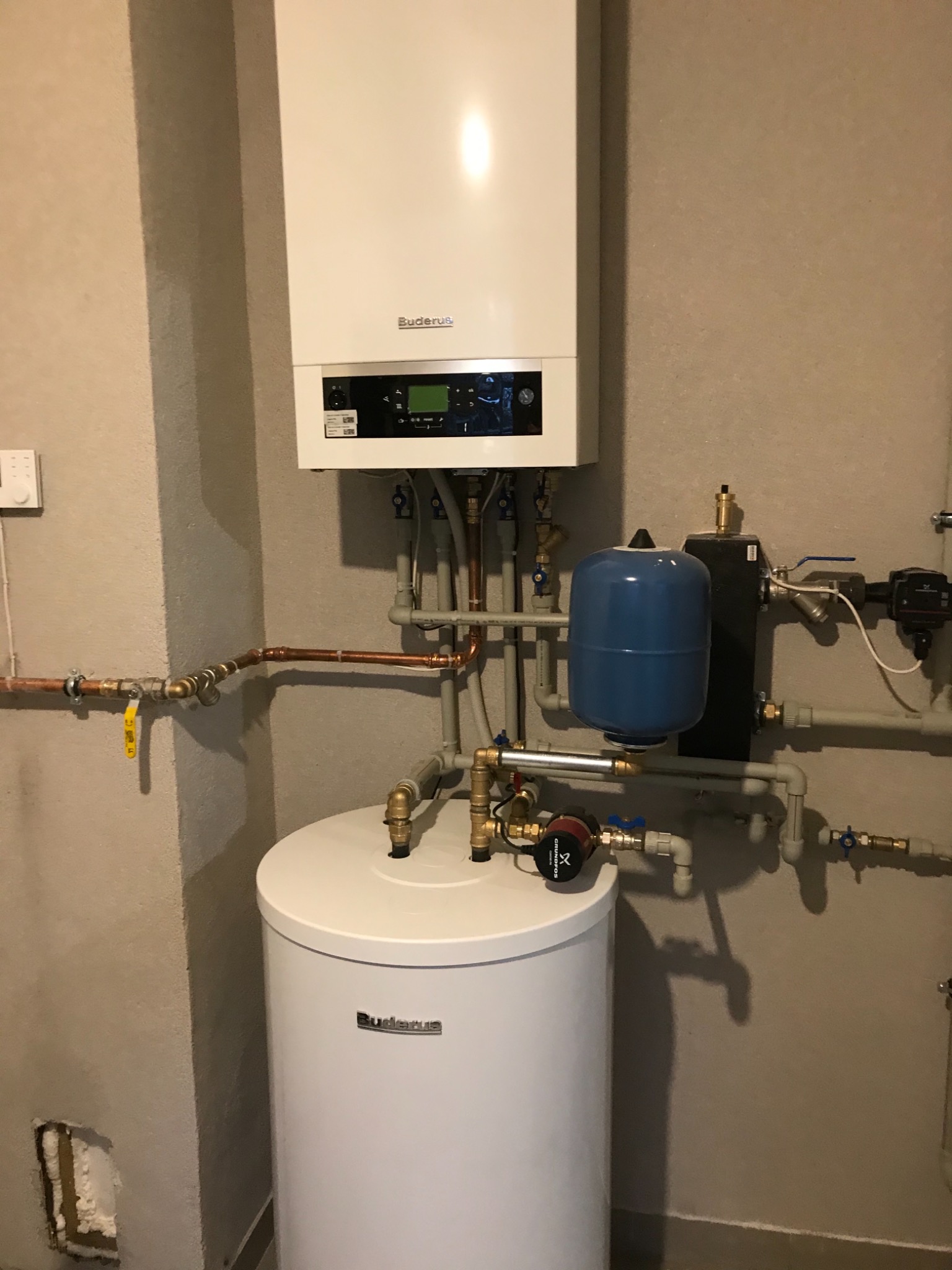 Biały piec gazowy Buderus zamontowany na ścianie, połączony rurami miedzianymi i plastikowymi z białym zasobnikiem ciepłej wody i niebieskim naczyniem przeponowym, widoczne zawory i pompa obiegowa...