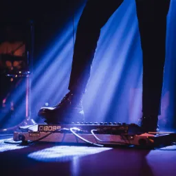 Stopa w skórzanym bucie naciska na efekt gitarowy BOSS ME-90 w niebieskim świetle scenicznym podczas występu.