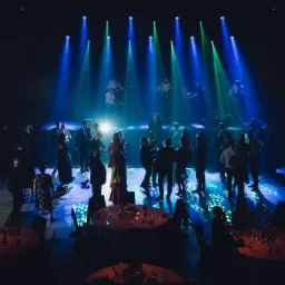 Widok z góry na salę balową z bawiącymi się gośćmi i zespołem grającym na scenie, oświetloną niebieskimi i zielonymi reflektorami.