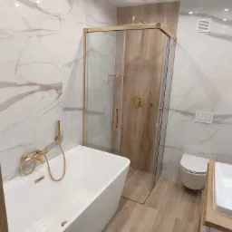 Nowoczesna łazienka z białymi marmurowymi płytkami, złotą armaturą i kabiną prysznicową z drewnianym panelem, widok na wannę, toaletę i umywalkę.