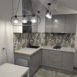 Nowoczesna kuchnia w odcieniach szarości z blatem imitującym jasny kamień, ozdobiona płytkami z motywem liści, oświetlona wiszącymi lampami z okrągłymi kloszami.