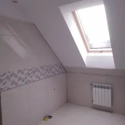 Wykończona łazienka na poddaszu z oknem dachowym, jasne płytki z dekoracyjnym pasem w zygzaki, biały grzejnik, widoczne ślady po pracach wykończeniowych.