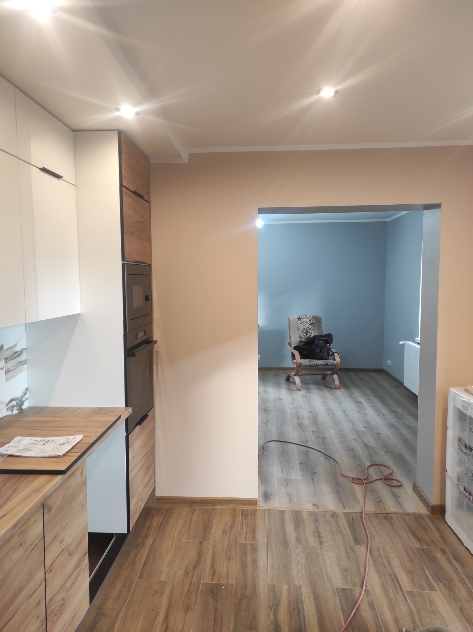 Nowoczesna kuchnia z drewnianymi i białymi szafkami, widok na pokój z szarą ścianą i krzesłem, podłoga drewnopodobna, widoczny przewód odkurzacza.