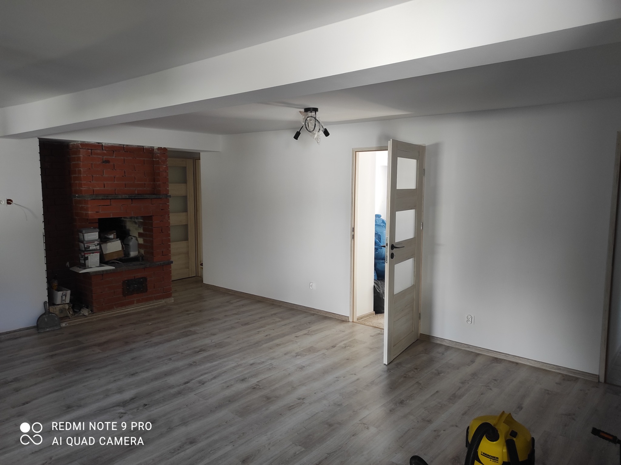 Wnętrze po odświeżeniu: pomalowane na biało ściany, położone panele podłogowe w odcieniach szarości, widoczny ceglany kominek i częściowo zamontowana lampa w folii ochronnej.