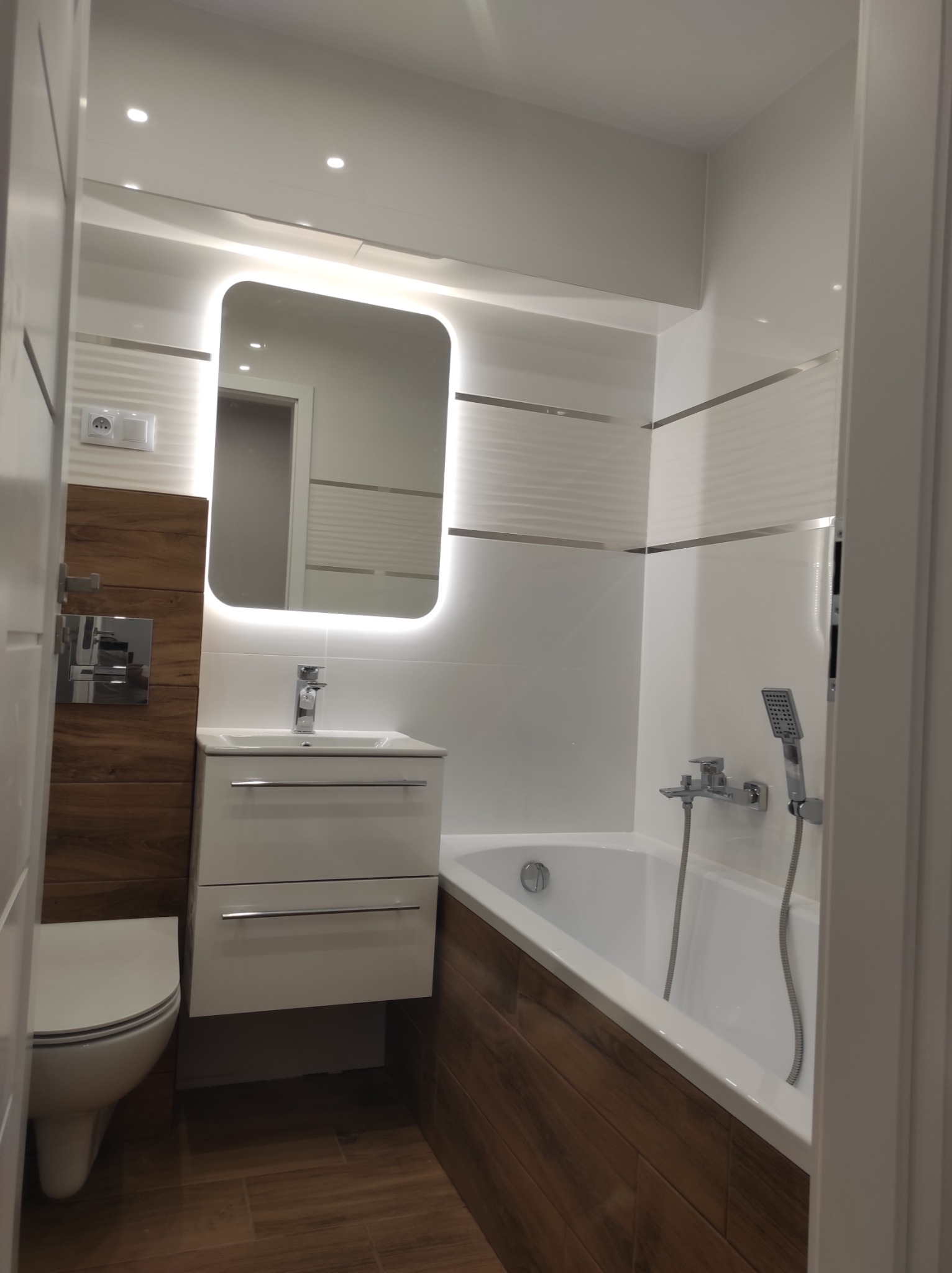 Nowoczesna łazienka po remoncie: białe płytki z akcentami drewna, podświetlane lustro, umywalka z szafką i wanna z prysznicem.