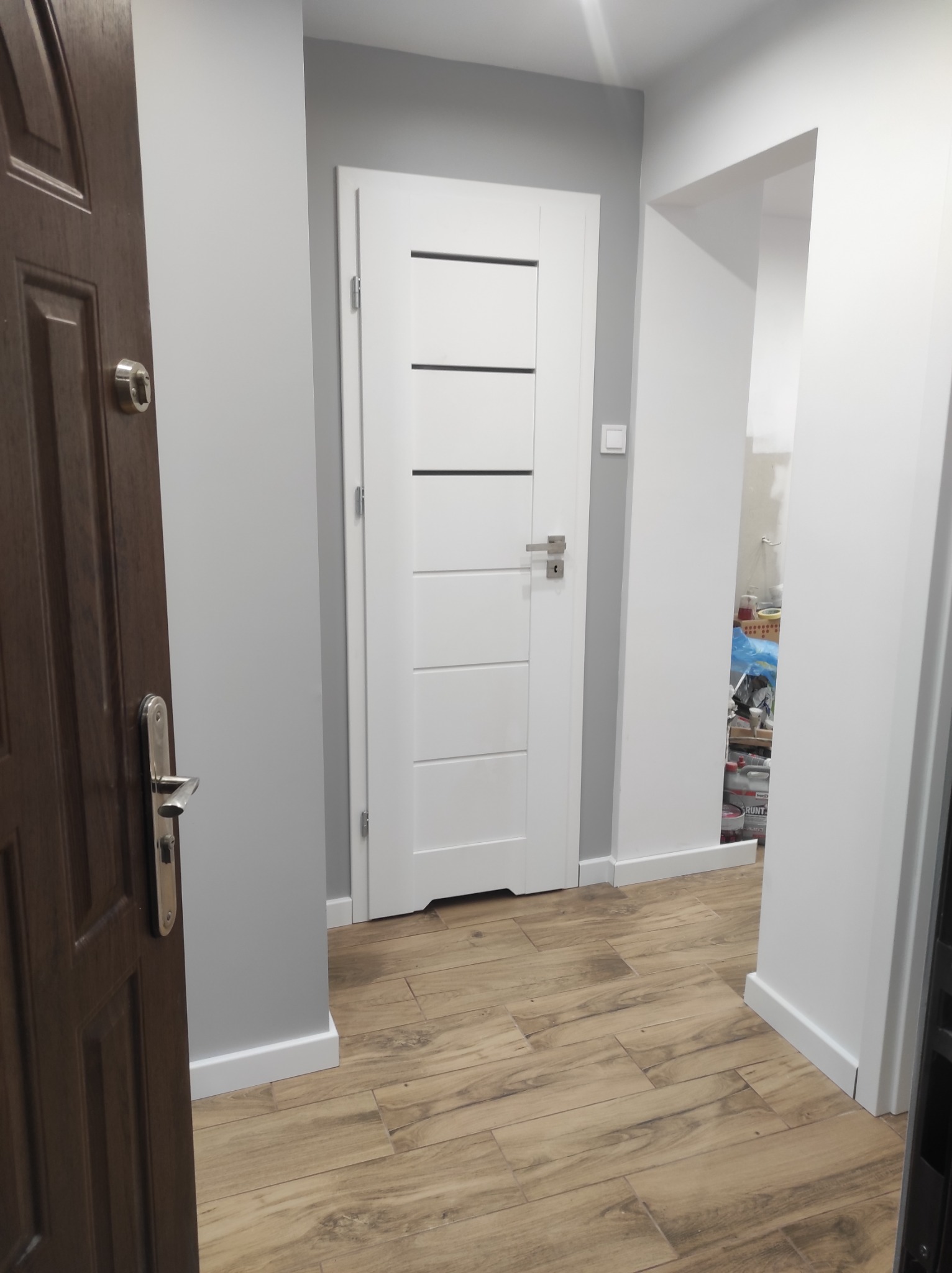 Wykończony korytarz z brązowymi panelami podłogowymi imitującymi drewno, szarą ścianą i dwiema parami drzwi: ciemnobrązowe wejściowe i białe wewnętrzne z poziomymi przeszkleniami.