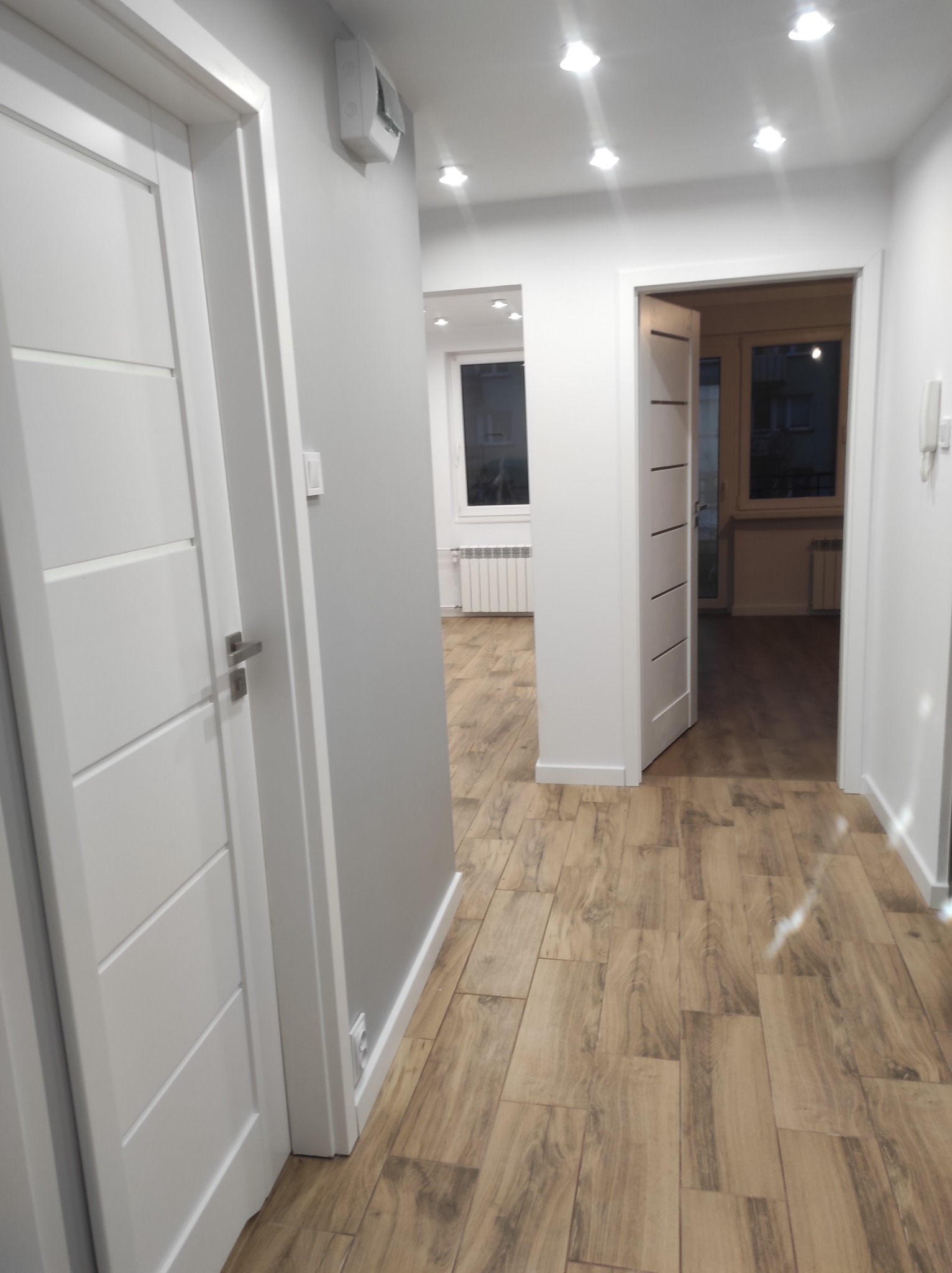 Jasny korytarz z drewnopodobną podłogą w odcieniach beżu i brązu, białe drzwi z poziomymi frezowaniami i szare ściany, widok na pokoje z białymi grzejnikami.