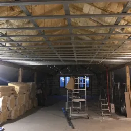 Poddasze w trakcie ocieplania, widoczne metalowe profile sufitowe i warstwa izolacyjna, stos wełny mineralnej przy ścianie, oraz rozstawione drabiny.