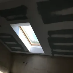 Poddasze w trakcie prac wykończeniowych: widoczne łączenia płyt gipsowo-kartonowych na skosach, przygotowane do szpachlowania, z oknem dachowym wpuszczającym światło dzienne.