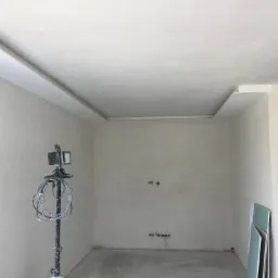 Pokój w trakcie wykańczania: świeżo położone gładzie na ścianach i suficie z dekoracyjnym obramowaniem, widoczna lampa budowlana na statywie i płyty karton-gips oparte o ścianę.