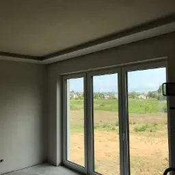 Wnętrze pokoju w stanie deweloperskim z dużym, trzyskrzydłowym oknem balkonowym z widokiem na pole i zabudowania. Ściany przygotowane pod malowanie, sufit z dekoracyjnym obramowaniem.