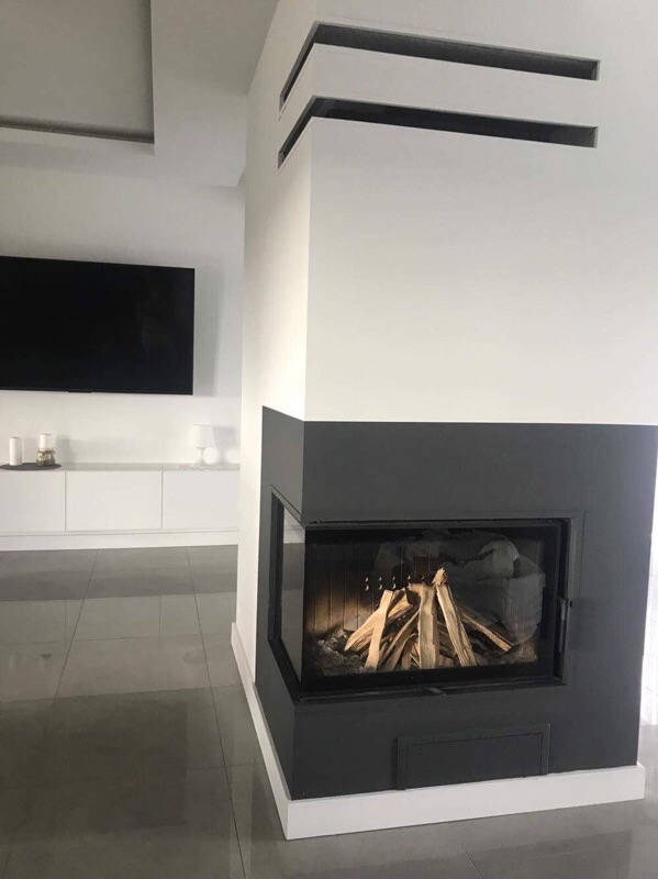 Nowoczesny kominek narożny z czarną obudową i panoramiczną szybą, w minimalistycznym salonie z szarą podłogą i białymi meblami.