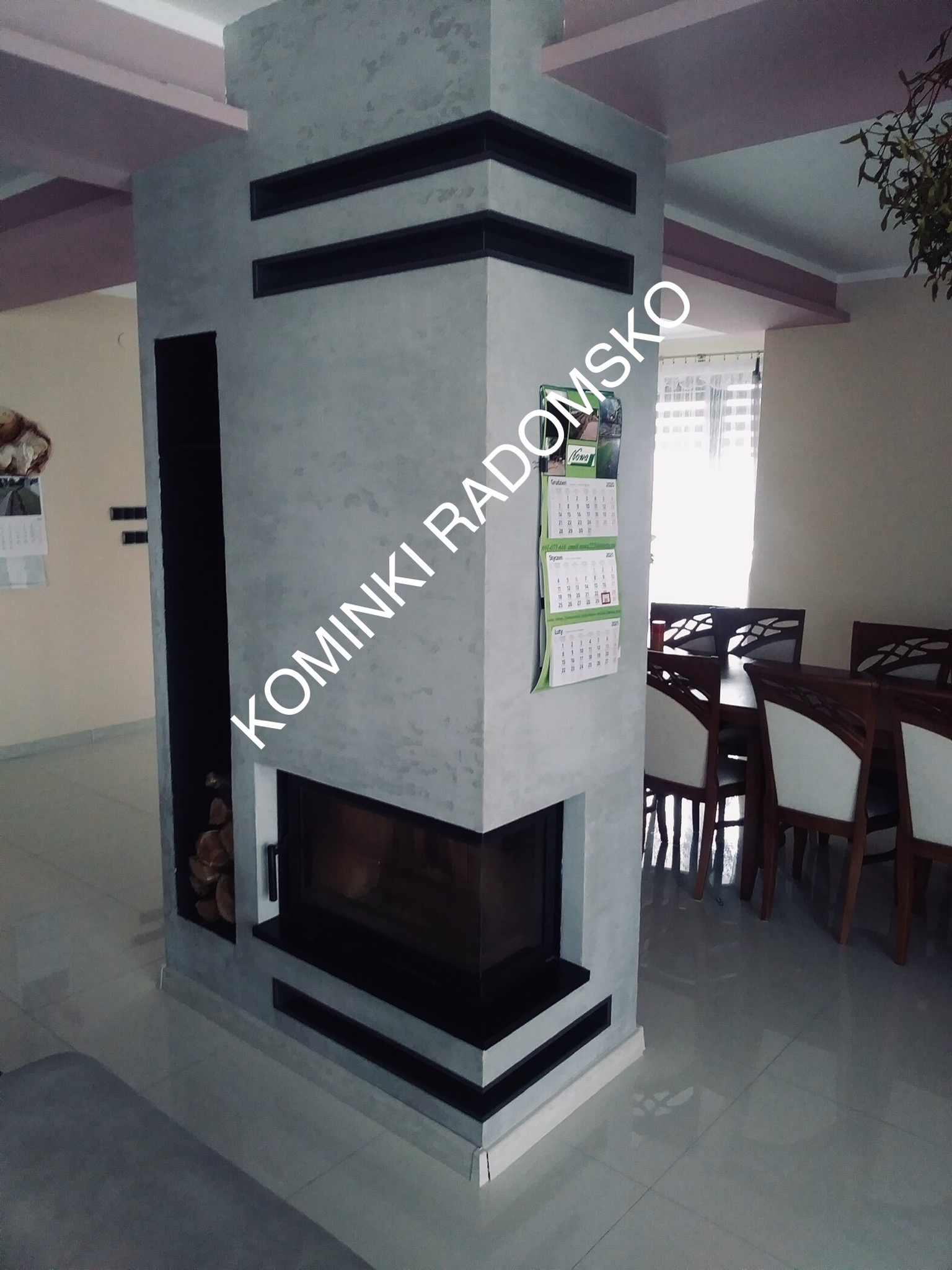 Nowoczesny kominek narożny z szarą strukturą tynku, czarnymi wstawkami i wnęką na drewno, widoczny w tle stół z krzesłami w jasnym wnętrzu.