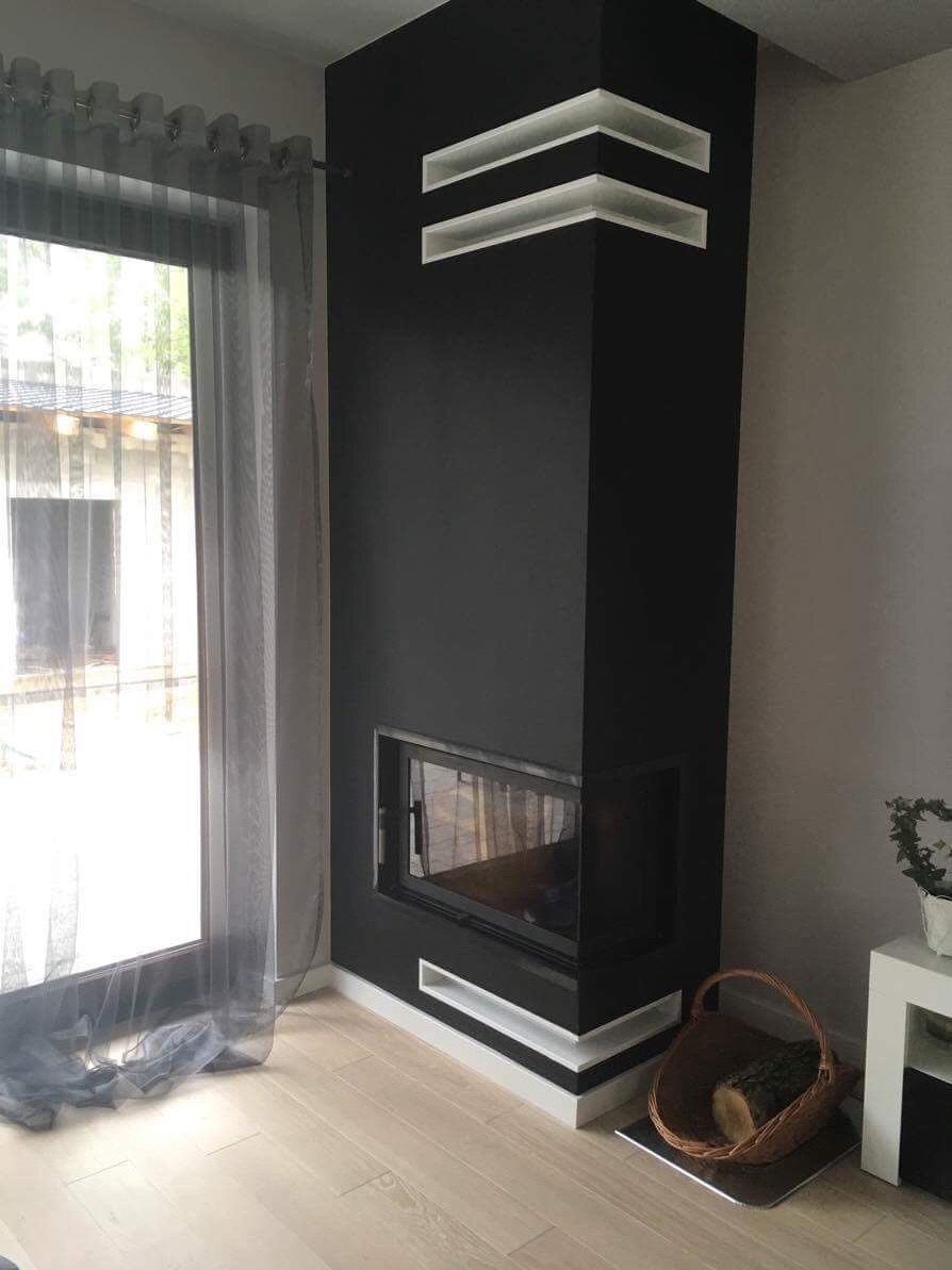 Nowoczesny, narożny kominek w kolorze czarnym z białymi, minimalistycznymi wnękami, ustawiony na jasnej podłodze z parkietu, obok okna z szarą zasłoną i koszem na drewno.