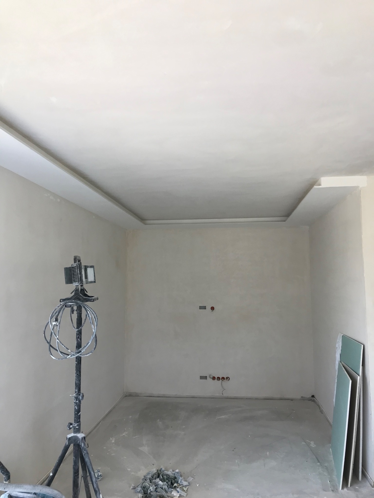 Pokój w trakcie wykańczania: świeżo położone gładzie na ścianach i suficie z dekoracyjnym obramowaniem, widoczna lampa budowlana na statywie i płyty karton-gips oparte o ścianę.