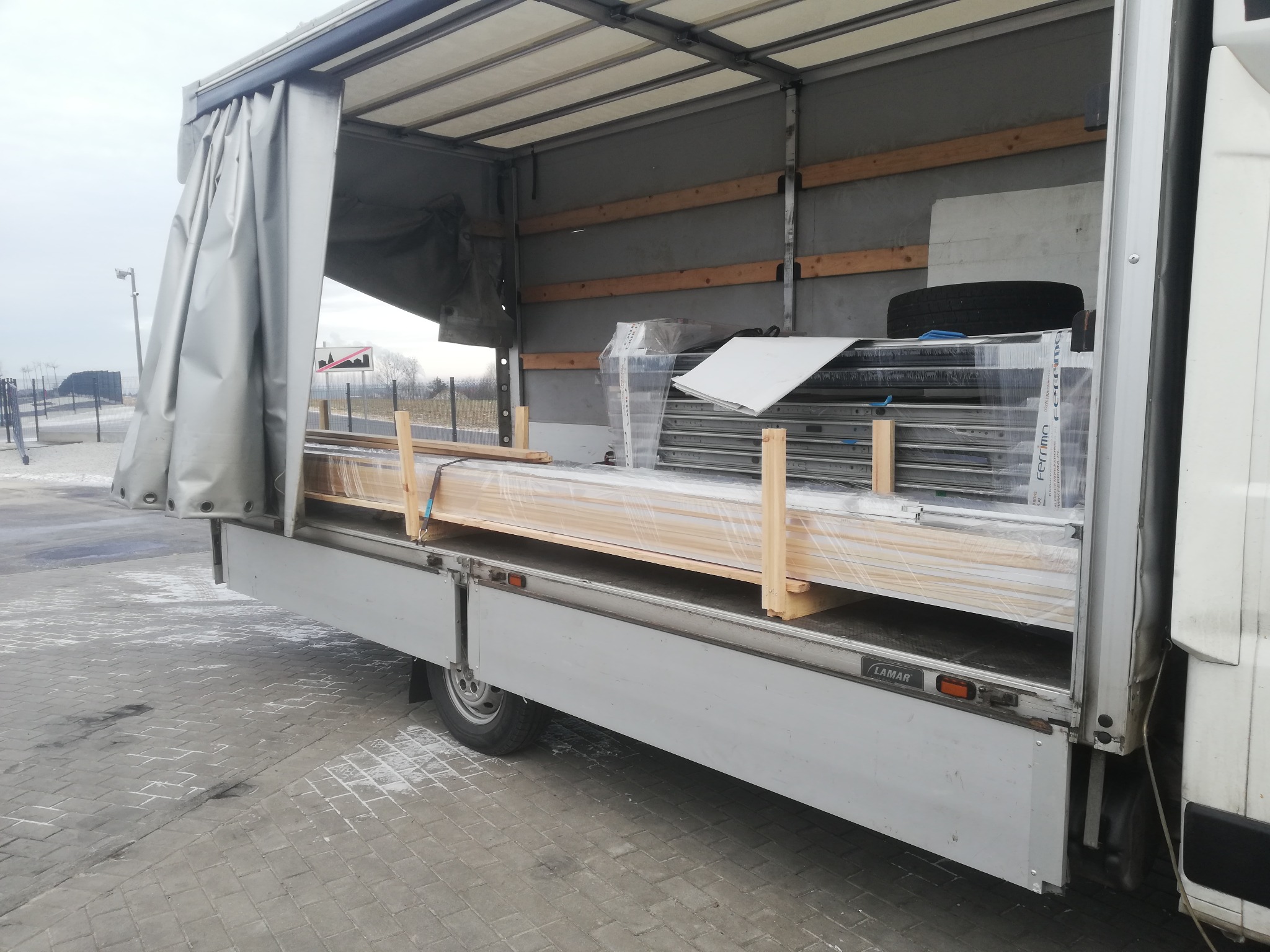 Otwarta paka busa z częściowo odsłoniętą plandeką, załadowana elementami okiennymi zabezpieczonymi drewnianymi belkami i folią stretch.