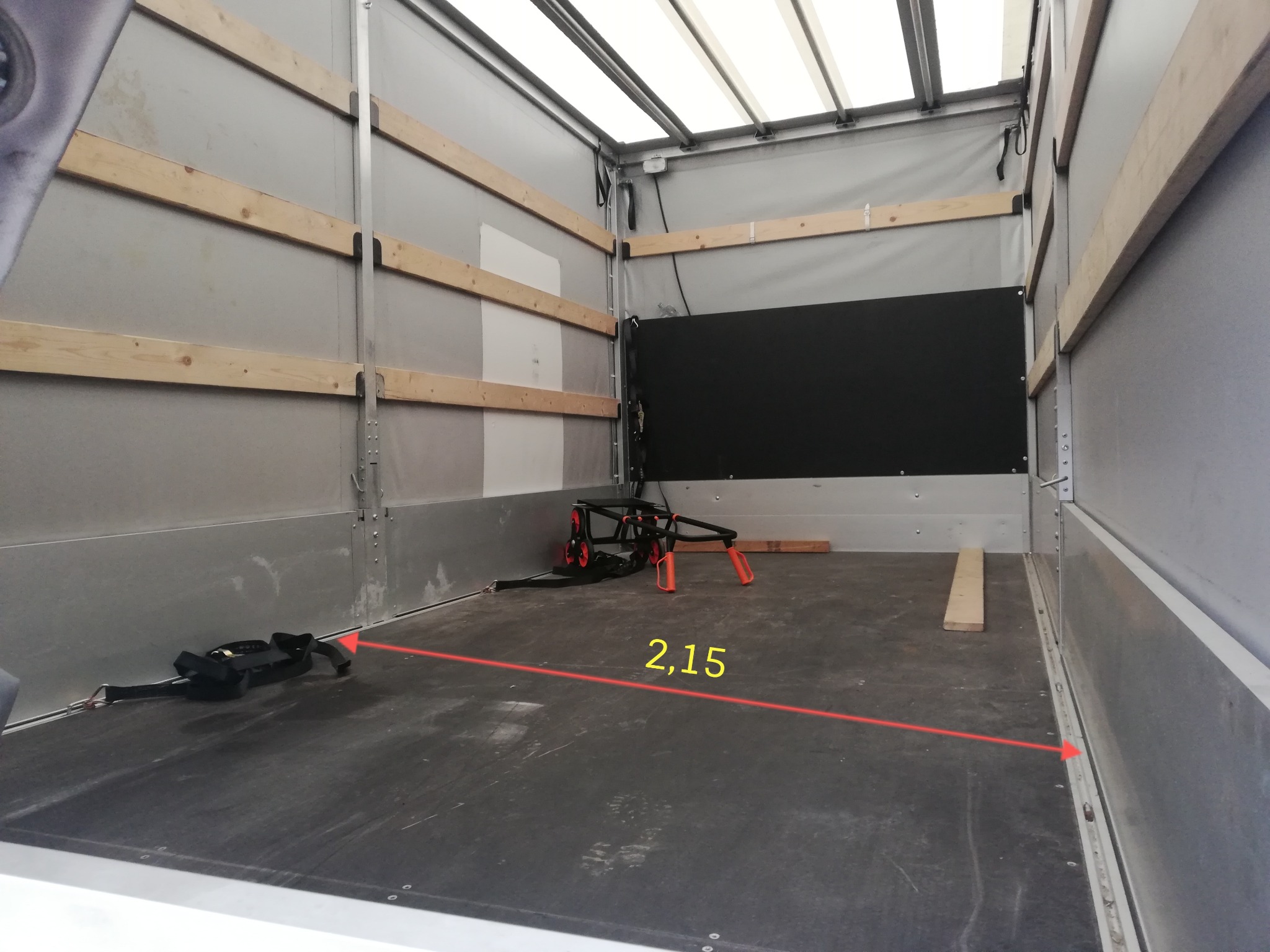Wnętrze pustej paki dostawczego busa z drewnianymi listwami mocującymi na ścianach, czarną przegrodą z tyłu i wózkiem transportowym na podłodze; widoczna linia pomiarowa 2,15 m.