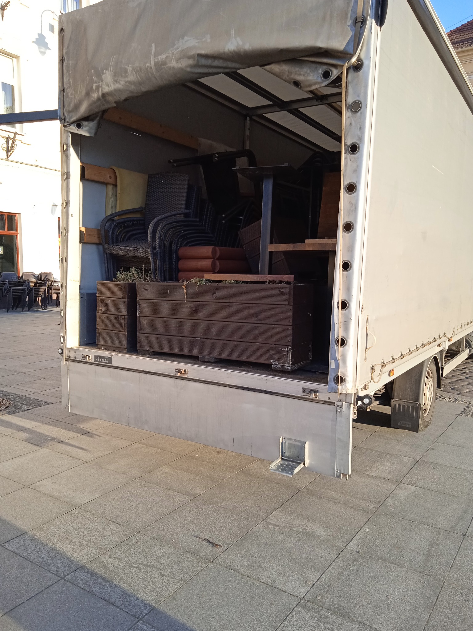 Otwarta paka busa wypełniona stosem wiklinowych krzeseł, drewnianymi donicami z roślinami i elementami stołów, widok z tyłu na chodnik z kostki.