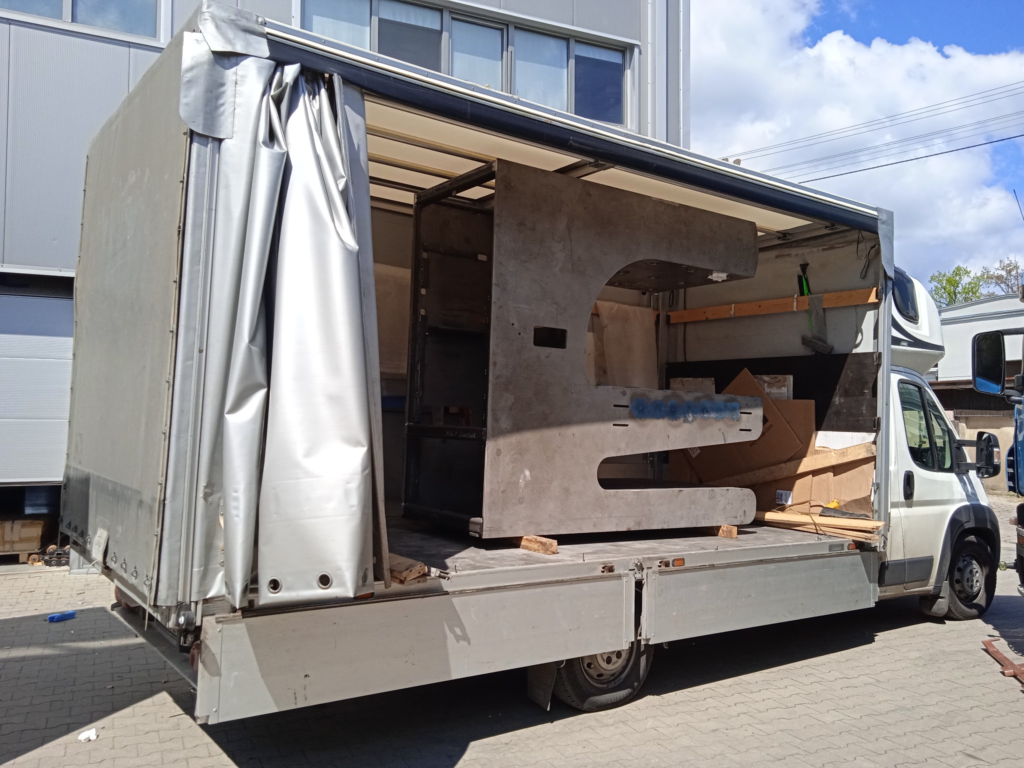 Otwarta paka busa z metalową konstrukcją o nietypowym kształcie, zabezpieczoną elementami drewnianymi i kartonami, widok z boku na tle budynku przemysłowego i fragmentu nieba.