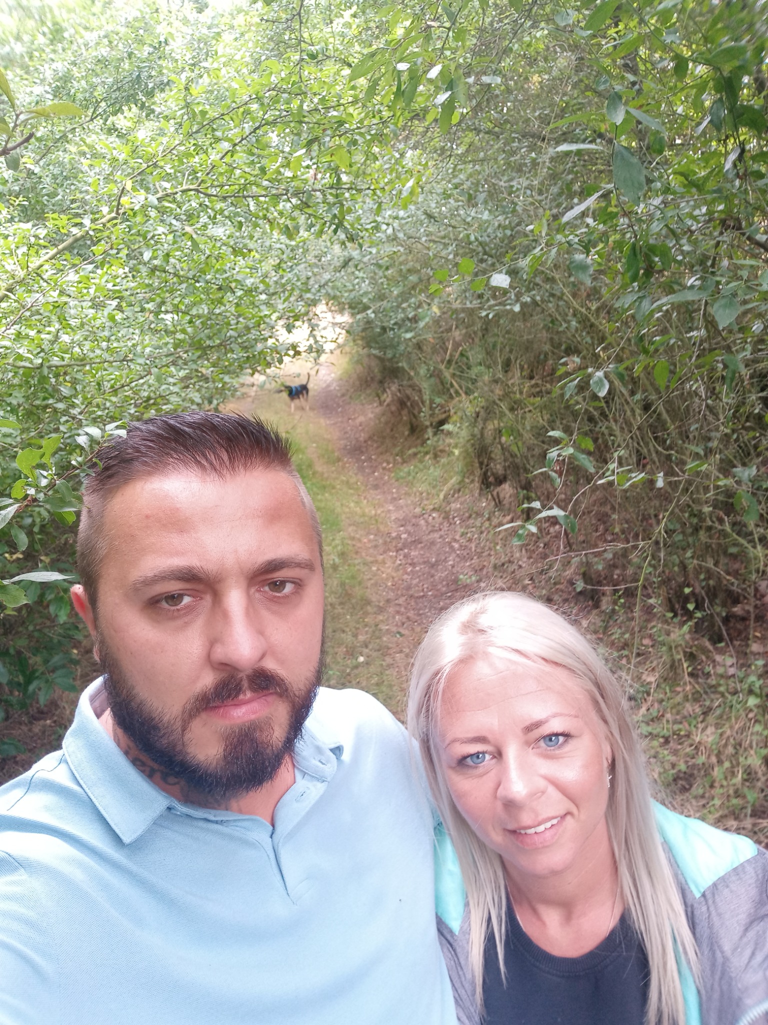 Selfie pary na leśnej ścieżce z psem w oddali, zarośniętej zielenią.