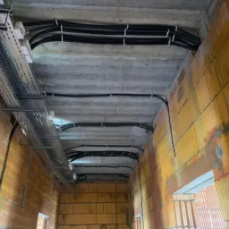 Kable elektryczne w peszlach i metalowe korytka kablowe zamontowane pod betonowym sufitem w surowym budynku z cegły dziurawki.